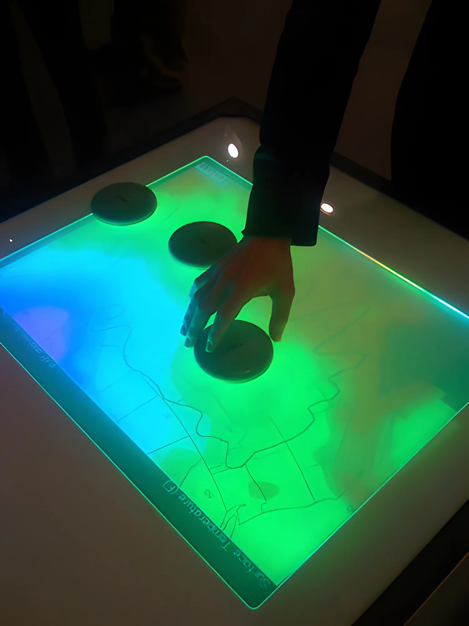 Looking down on the darkened table, one hand rests on the nearest of three dark pucks; the map beneath glows green on the right and blue-cyan on the left.
