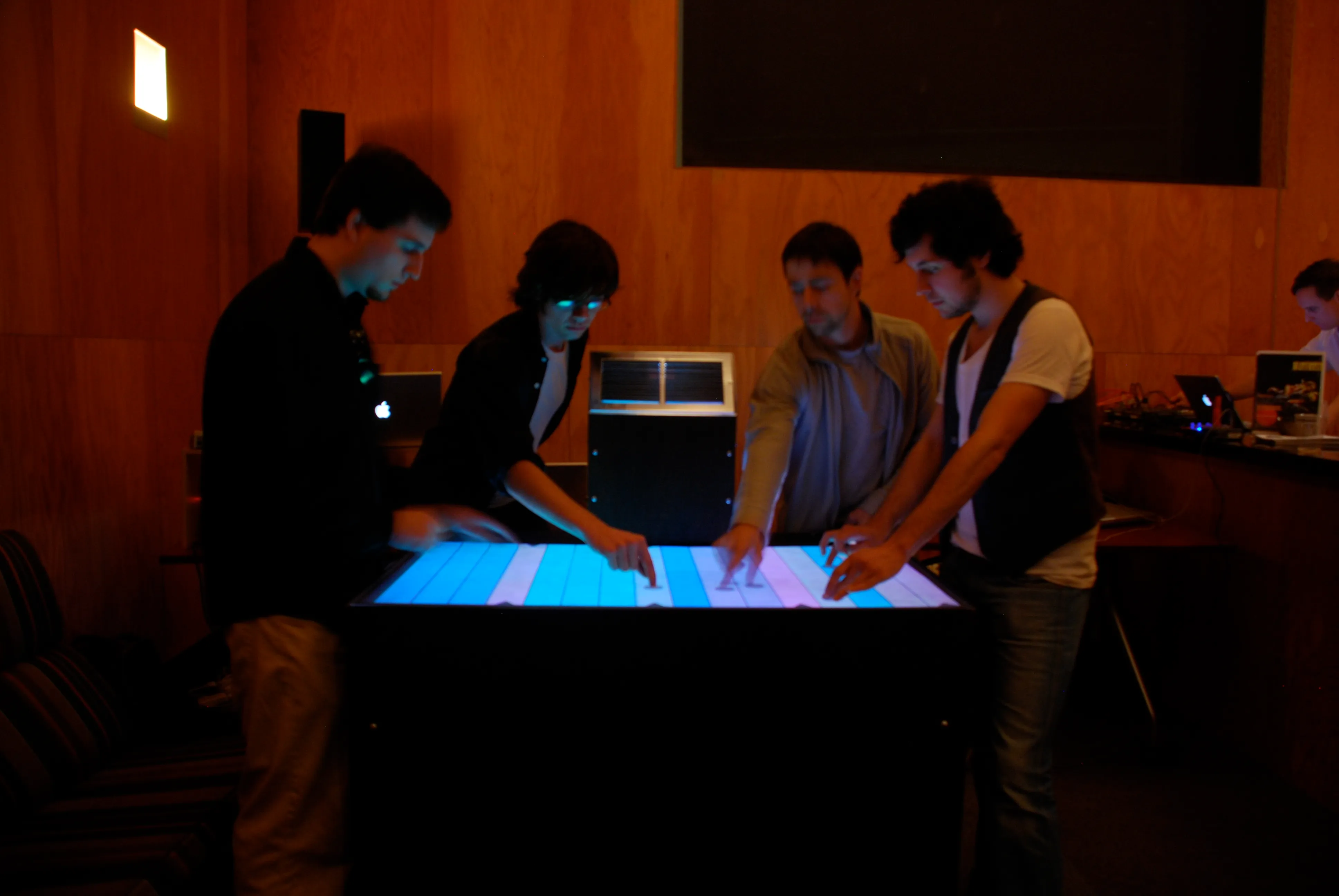 Four players stand around the table in a wood-panelled room, hands on a row of vertical blue, white and pink bands lighting the surface.