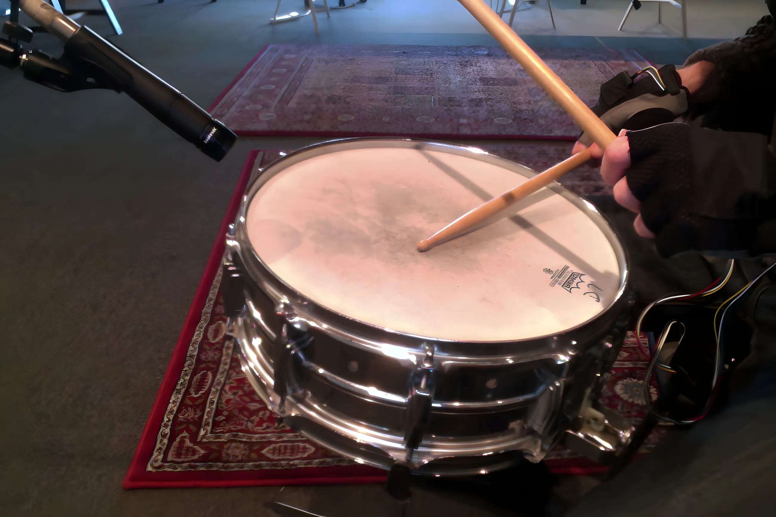 Over-the-shoulder view of a gloved hand striking a chrome snare with wires strapped along the forearm, mic overhead
