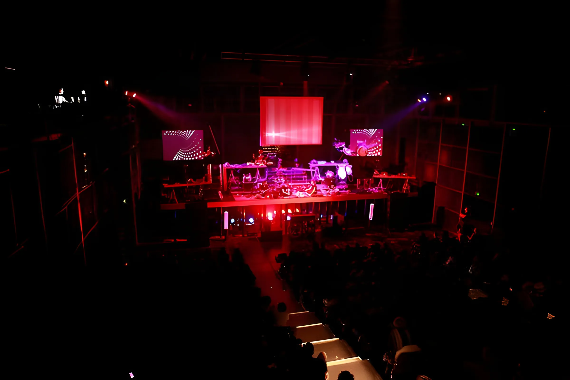 Red-lit hall from the balcony, silhouetted audience below a stage of machines and three glowing screens.