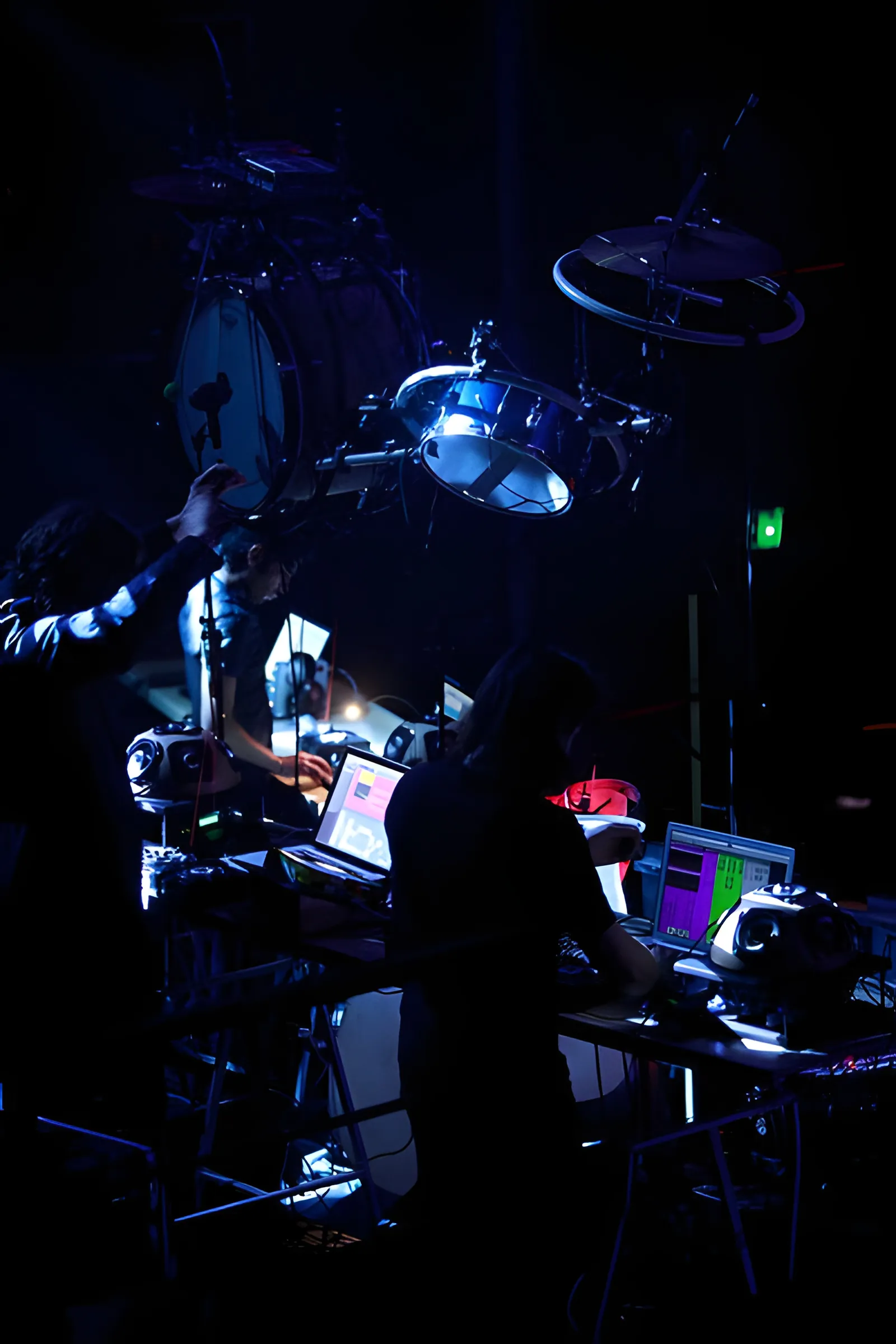 A performer reaching up to a drum kit suspended overhead while two others work at glowing laptops.